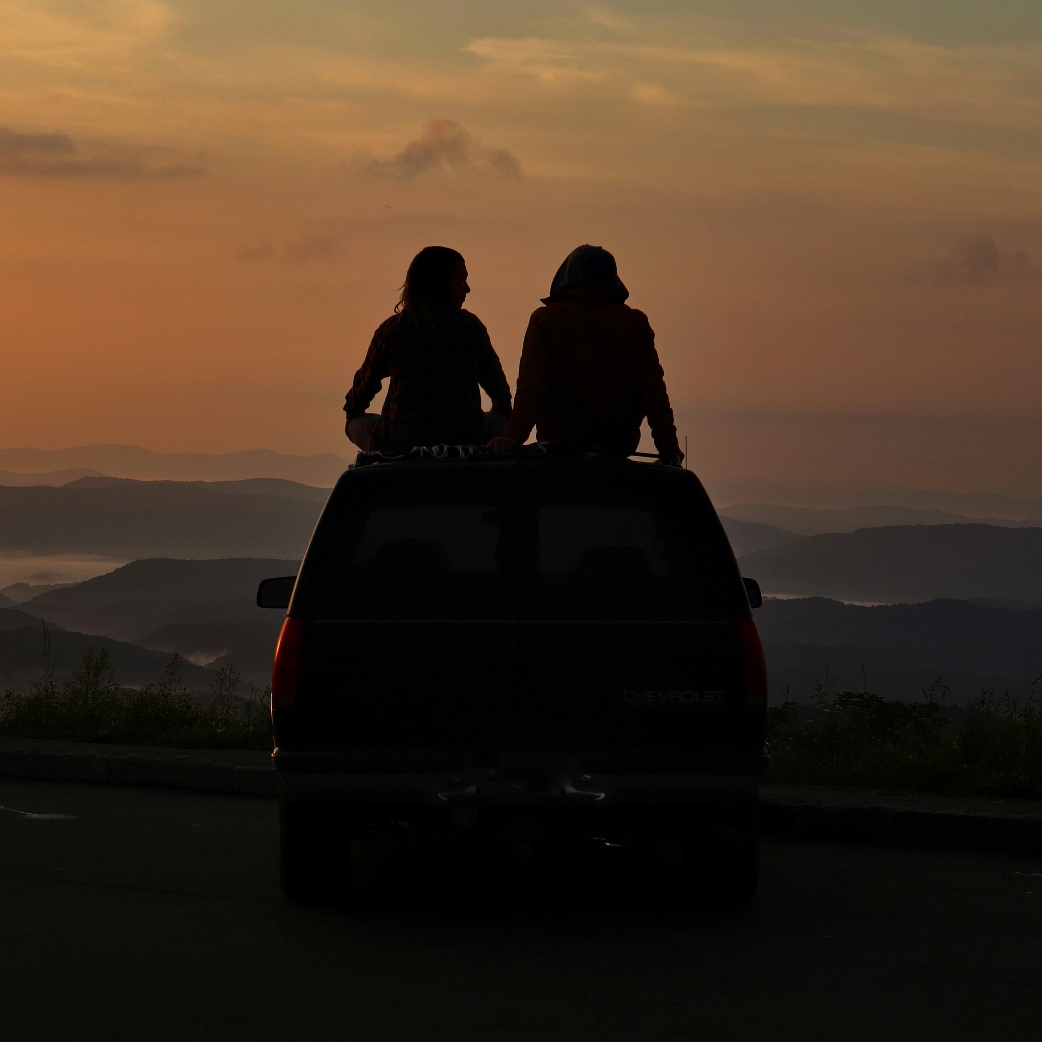 Duas pessoas sentadas no teto de uma caminhonete observando o pôr do sol sobre montanhas enevoadas no Brasil